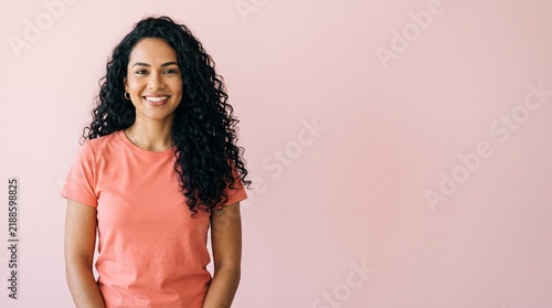 Smiling woman with curly dark hair wearing a coral t-shirt against a soft pink background