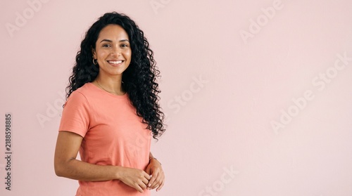 Smiling young woman with long curly dark hair wearing a coral t-shirt against a soft pink background