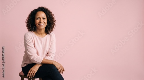 Smiling middle aged woman with curly hair sitting on a stool against a pink background