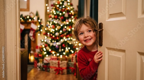 Joyful young child peeking around a door with a decorated Christmas tree and presents in the background