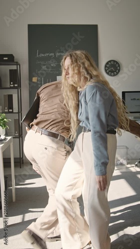 Vertical video of Asian man and Caucasian woman shaking hands and dancing playfully with folder in office. Footage fits corporate culture campaigns and employee satisfaction promotions