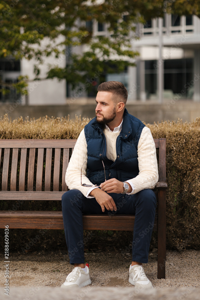 Fototapeta premium Taking a moment to relax outdoors, a casually dressed man sits comfortably in a stylish vest surrounded by autumn foliage. The cozy setting, natural light and warm colors create a calm seasonal atmosp