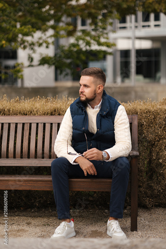 Taking a moment to relax outdoors, a casually dressed man sits comfortably in a stylish vest surrounded by autumn foliage. The cozy setting, natural light and warm colors create a calm seasonal atmosp