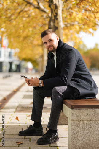 Stylish businessman in an elegant coat sitting outdoors, talking on his phone on an autumn day. Warm fall colors, modern urban setting and natural light.
