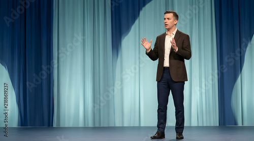 Professional business speaker, delivering persuasive keynote address with hand gestures, dressed in smart casual blazer, standing before blue curtain stage, corporate conference event marketing.
