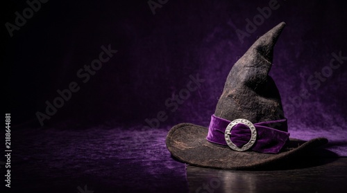 A spooky witch hat with a purple velvet ribbon and silver buckle sits on a dark wooden surface against a deep purple background