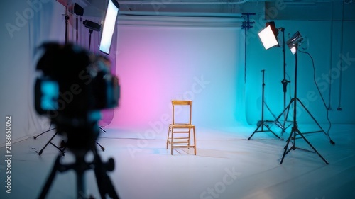 Professional photography studio with lighting equipment, empty wooden chair on white backdrop, modern film set with neon pink and blue lights