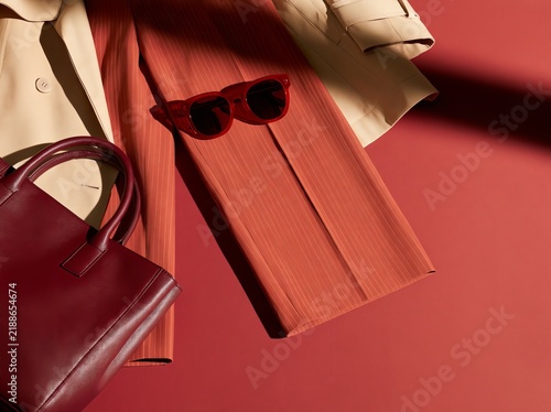 Tailored workwear accessories including a beige trench coat, red pinstripe trousers, red sunglasses, and a burgundy handbag arranged on a flat lay against a red background