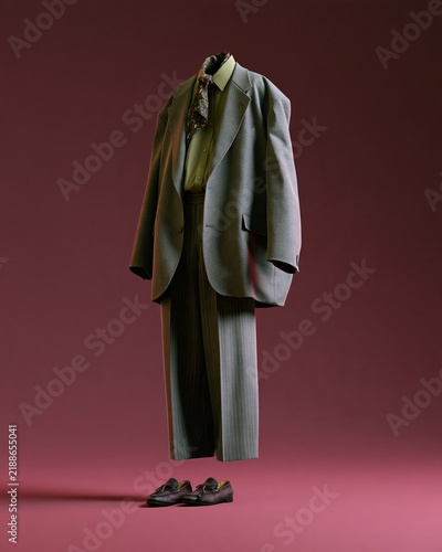 Floating ghost mannequin displaying oversized gray suit with matching trousers, green shirt, and patterned scarf against a solid burgundy background