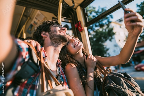 Young couple taking selfie with smartphone on public bus during travel adventure