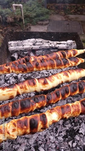 Close-up of hands turning skewers with sulguni cheese wrapped in golden puff pastry dough, baking on charcoal grill with smoke and flame
