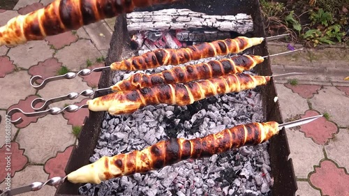Close-up of hands turning skewers with sulguni cheese wrapped in golden puff pastry dough, baking on charcoal grill with smoke and flame