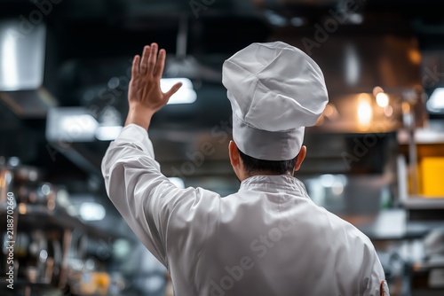 Back view of a professional chef in uniform raising hand in a busy commercial kitchen, gesturing or signaling while working in a modern restaurant environment.