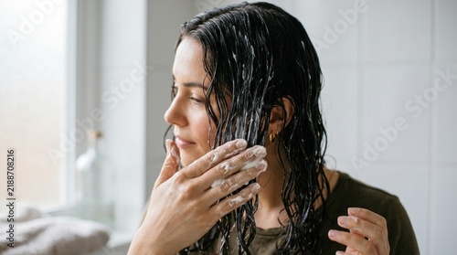 Woman applying conditioner to wet dark hair in a bathroom.