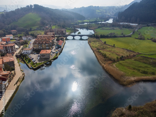 Aerial view of the Asón River with cloud reflections, the town of Limpias and its promenade, surrounded by green fields and hills. Limpias, Cantabria, Spain.
