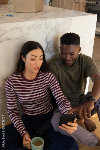 Diverse couple sitting on kitchen floor leaning against island holding phone and mug, box and keys