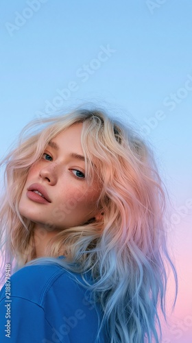 Close-up fashion portrait of a woman with long wavy pastel pink blonde hair and blue clothing against a dreamy blue and pink gradient background Theme of expressive hair beauty