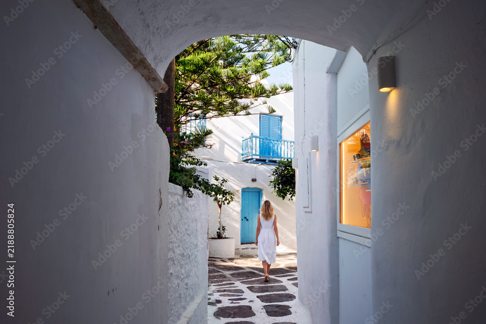 Fototapeta premium A young woman walks down a narrow whitewashed arched alleyway with colorful doorways and shutters in the old town center of Mykonos, Greece.