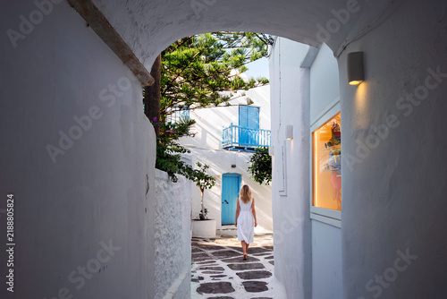 A young woman walks down a narrow whitewashed arched alleyway with colorful doorways and shutters in the old town center of Mykonos, Greece.