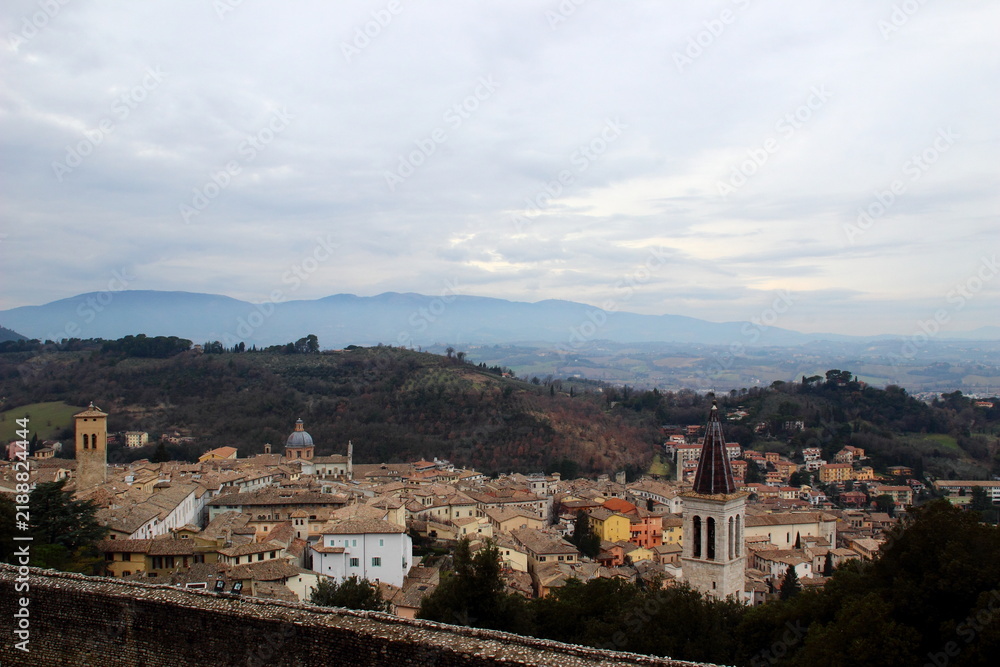 Fototapeta premium Scenic view of Spoleto with terracotta rooftops and church towers in front of thickets, hills, distant mountains, creating a particular, longing atmosphere, under a cloudy, airy, spiritual sky