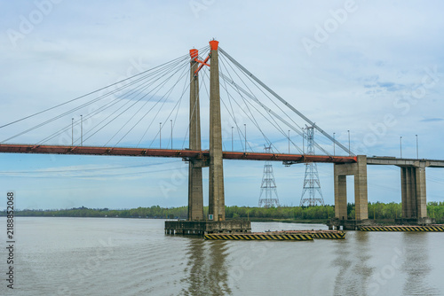 Wide side view of a large cable-stayed suspension bridge spanning over a calm river, featuring tall concrete towers, red roadway deck, and electric power line pylons nearby