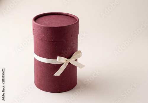 A maroon cylindrical gift box tied with a white ribbon and bow. The box has a textured surface and a lid on top.