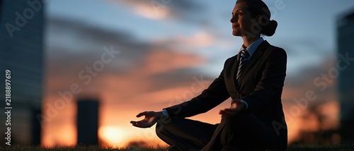 Businesswoman in formal suit meditating peacefully outdoors at sunset with city skyline background