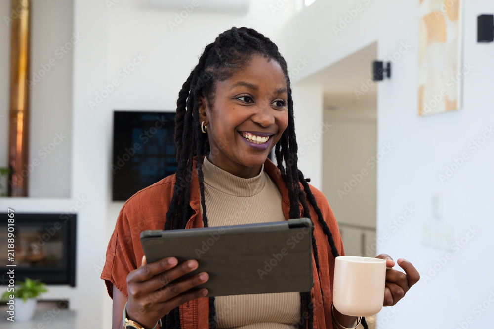 Fototapeta premium African American woman smiling, holding tablet and mug in bright living room, wearing rust jacket