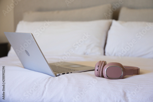 Silver laptop is sitting open on white bed, matte pink headphones are resting near headboard