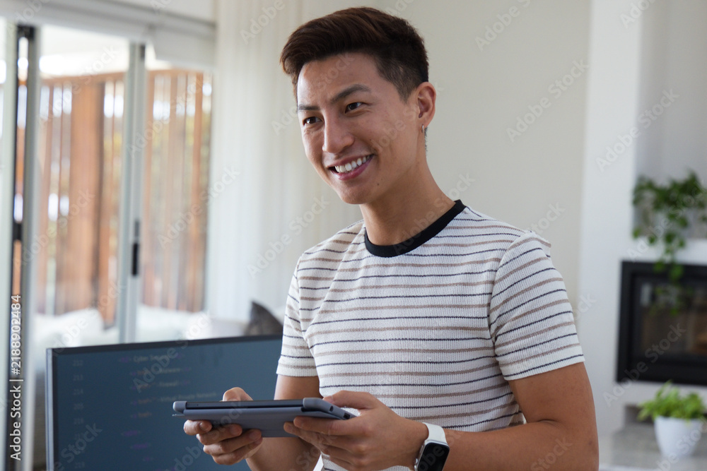 Obraz premium Mid-adult Asian man standing holding tablet with smartwatch in living room monitor showing code