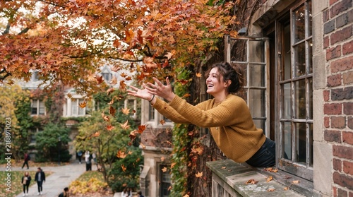 Joyful young woman throwing autumn leaves from a window on a university campus during fall season