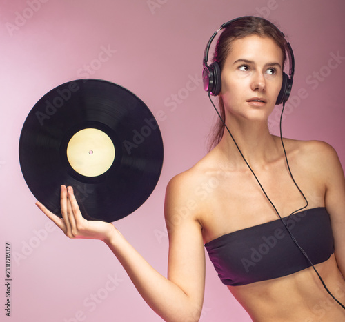 A young female student holds a vinyl record and listens to music on headphones against a white background.