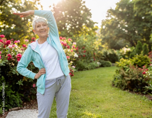 Active senior woman exercising and stretching in a green park with flowers during morning sunlight