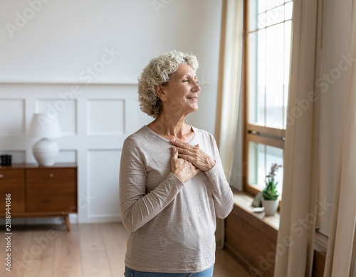 Peaceful senior woman expressing gratitude with hands on heart in sunlit room