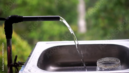 Clean water flowing from a pipe into a stainless steel sink in an outdoor area against a lush green foliage backdrop.