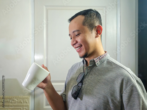 Canvastavla Smiling Asian man holding white coffee mug looking happy indoors during morning