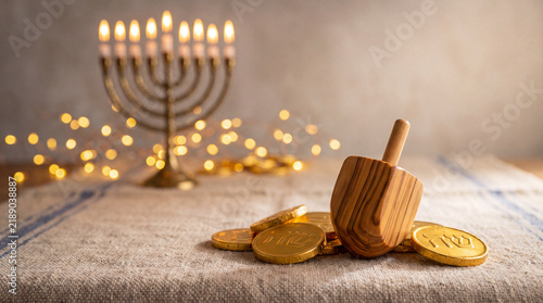 A traditional wooden dreidel rests on gold chocolate coins, with a blurred Hanukkah menorah and festive fairy lights glowing in the background.