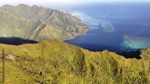 Aerial view circling high volcanic ridges to reveal Opunohu Bay, featuring a shallow coral reef lagoon, anchored sailboats, and the deep blue Pacific ocean