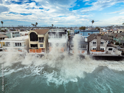 Large waves crash against oceanfront homes during heavy surf, sending spray above seawalls and highlighting the risks of coastal flooding and erosion