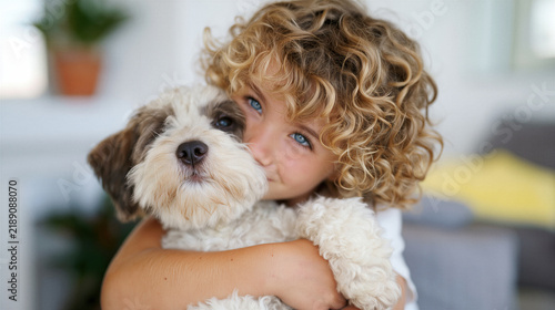 Curly haired child with blue eyes hugging fluffy white and brown dog indoors warmly