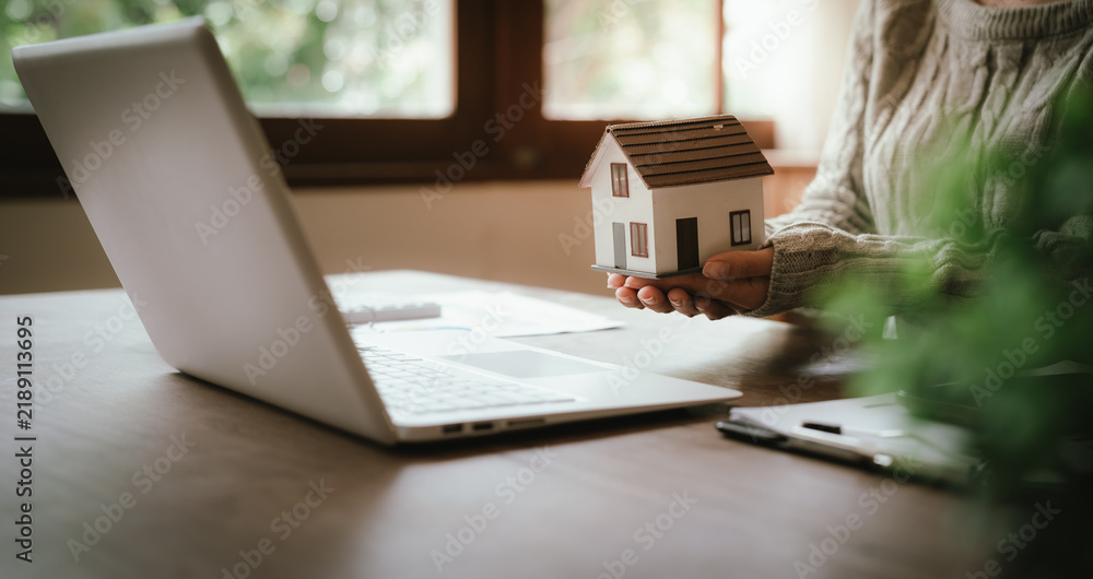 Fototapeta premium Real estate professional holding a model house at a desk with property documents, representing home ownership, mortgage, investment, and property transactions.
