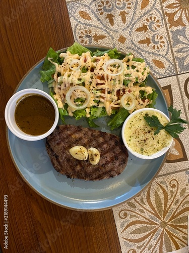 Foto Grilled beef steak topped with garlic butter served with fresh salad, creamy side, and gravy sauce on a blue plate over a patterned table
