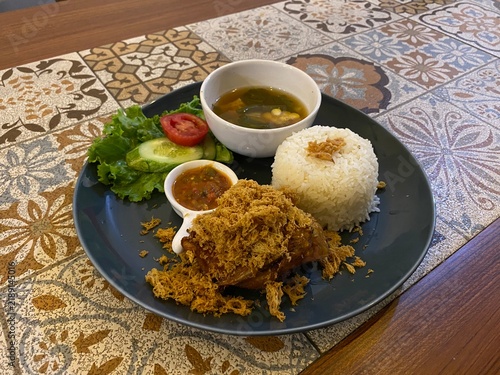 Konstfotografi Indonesian fried chicken meal served with steamed rice, soup, chili paste, cucumber, and lettuce on a dark plate on a wooden table