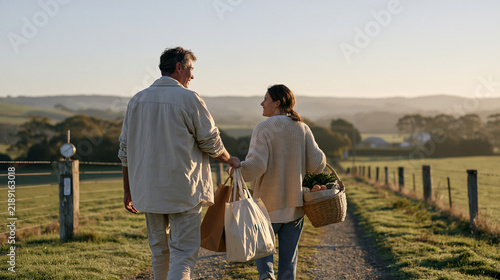 Occupational therapist stepping back on a misty early morning farm path to assess a patient carrying reusable grocery bags from a market stall, a barometric gauge on a nearby post