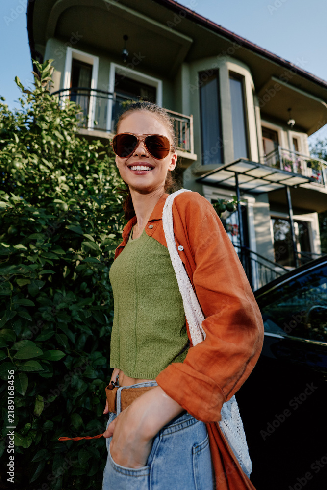 Obraz premium Smiling young woman in sunglasses and orange shirt poses near greenery and a house on a sunny day. Casual summer lifestyle, confident happy mood, street fashion portrait concept.