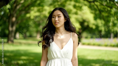 A young woman with flowing dark hair walks gracefully through a sunlit park. She's wearing a white dress