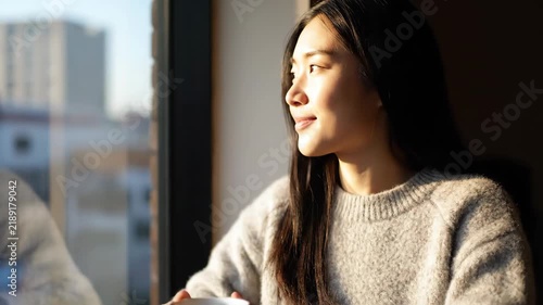 A woman gazes out a window with a cup, lit by sunlight, relaxed and serene, wearing a sweater