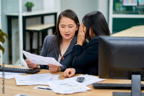 Asian businesswomen whispering confidential information and secret office gossip at desk during working hours. Corporate women discussing financial paper report.