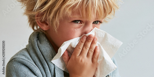A young boy with blond hair, wearing a robe, wipes his nose with a white tissue. He looks unwell, with his eyes partially closed and a concerned expression