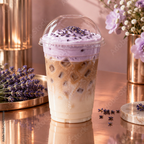 Iced lavender latte in a clear plastic cup with purple foam topping and dried lavender buds, set on a reflective copper surface with floral decor